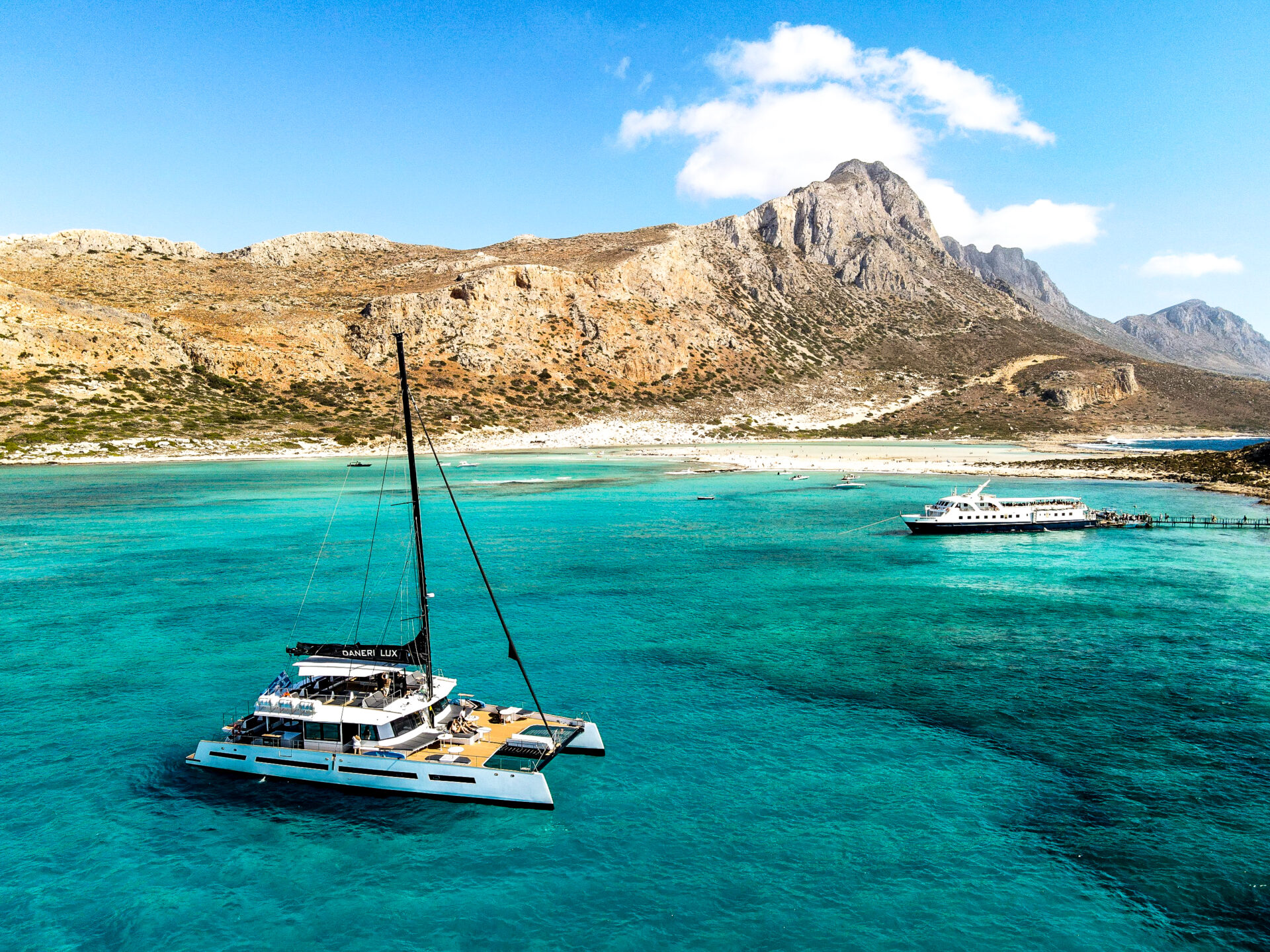A catamaran on turquoise water beside a mountainous Cretan coastline